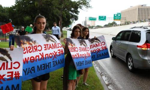 Young girls on the side of the road hold signs that read "617888九五至尊娱乐 Crisis, #ExxonKnew Make Them pay."